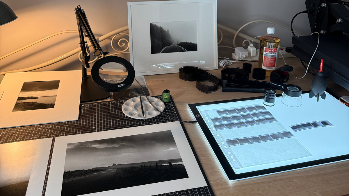 A black and white landscape photograph in a black frame hanging on a gray wall above a wooden desk with matted prints, a light box, magnifying glass, and other photography tools.